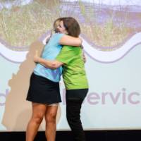 Teacher receives hug from Groundswell staff in front of projected "service award" background